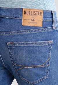 Jeans i denim med en ljusblå tvätt, med en läderlapp med "HOLLISTER"-logotyp, två bakfickor med orangea sömnadsdetaljer.