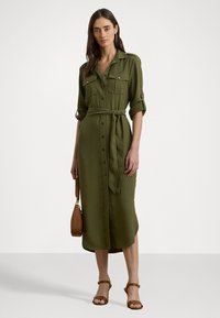 Φόρεμα σε ελιά πράσινο χρώμα με κουμπιά μπροστά, δύο τσέπες στο στήθος, ζώνη στη μέση και γυριστά μανίκια. Συνδυασμένο με καφέ σανδάλια με λουριά και καφέ τσάντα.