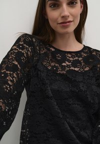Jauna moteris, vilkinti juodą gėlių nėriniuotą ilgomis rankovėmis marškinį, švelniai šypsosi prieš paprastą baltą foną.