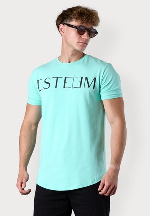 T-shirt mint verte à manches courtes avec le texte "ESTEEM" en lettres noires en gras, fabriqué en tissu coton lisse. Un short noir et des lunettes de soleil complètent le look.