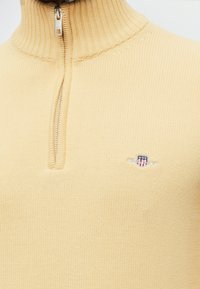 Pull tricoté couleur crème avec une fermeture éclair quart de tour et un petit logo écusson brodé sur la poitrine.
