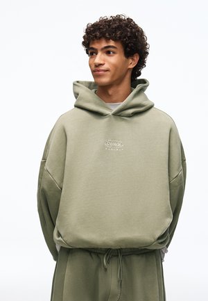 Giovane uomo con capelli ricci che indossa una felpa con cappuccio verde oliva e pantaloni della tuta abbinati, in piedi con le mani dietro la schiena, che guarda leggermente di lato.