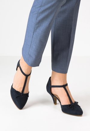 Navyblauwe suède hoge hakken met een spitse neus, T-band ontwerp en een strikaccent aan de voorkant. Gecombineerd met op maat gemaakte grijze broeken.