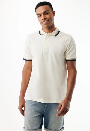 Young man smiling, wearing a white polo shirt with navy and beige trim, and light blue rolled-up denim shorts against a white background.
