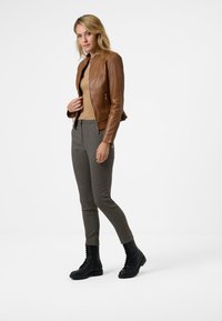 Veste en cuir marron ajustée sur un pull beige, assortie à un pantalon à carreaux et des bottes noires à lacets. La veste possède un col montant.