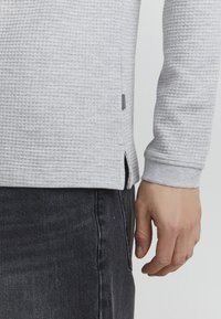Hellgraues, strukturiertes Langarmshirt mit gerippten Bündchen und seitlichen Schlitzen, kombiniert mit dunklen Jeans. Schwarzes Akzentetikett an der Seite des Shirts.