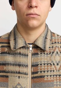 Wollmischung Reißverschlussjacke mit einem geometrischen Muster in Beige, Braun und Grau, mit Hemdkragen und sichtbarem Reißverschluss.