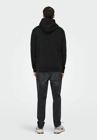 Zwarte hoodie met een relaxed fit, geribbelde manchetten en zoom. Gekozen in combinatie met donkergrijze jeans en lichtgekleurde sneakers, gezien van achteren.