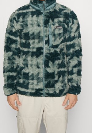 Mies, jolla on vihreä- ja harmaasävyinen maastokuvioitu fleece-takki, jossa on Columbian logo, yhdistettynä beigeruskeisiin housuihin yksiväristä taustaa vasten.