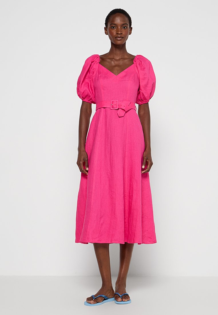 Frau in einem leuchtend pinken Midikleid mit Puffärmeln, tailliertem Bund und blauen Flip-Flops, die vor einem weißen Hintergrund steht.