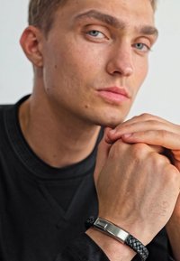 Woven black leather bracelet with a silver rectangular clasp, engraved with "BOSS." Model’s hands are positioned together, showcasing the accessory.
