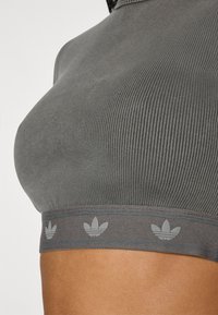 Šedý sportovní crop top s žebrovanou strukturou, hladkou látkou, kontrastním páskem s logem se třemi listy a přiléhavým atletickým střihem.