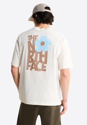 Person wearing a white T-shirt with brown "THE NORTH FACE" text and a large blue flower graphic on the back, paired with a light cap.
