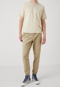 Beige katoenen t-shirt, tan elastische broek met cuffs, en blauwe hoge sneakers met witte rubberen zolen. Eenvoudig ontwerp met minimale branding.