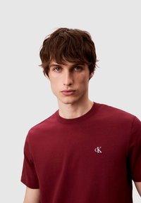 Bordeauxrotes Baumwoll-T-Shirt mit Rundhalsausschnitt, kurzen Ärmeln und einem kleinen weißen "cK"-Logo auf der linken Brust. Glatte Textur.