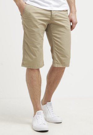 Mann trägt beige knielange Shorts, weiße Turnschuhe und ein weißes Hemd, steht mit entspannt hängenden Händen an den Seiten.