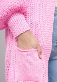 Cardigan rosa lavorato a maglia con una texture a costine, dotato di una grande tasca su un lato e maniche morbide e texturizzate.