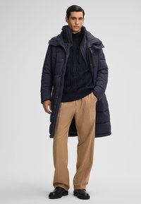 Man in een marineblauwe pufferjas over een zwarte kabelgebreide trui, beige broek en zwarte laarzen, staand tegen een effen grijze achtergrond.