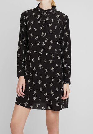 Femme portant une robe chemise noire à manches longues avec de petits motifs floraux blancs, debout devant un fond gris uni.