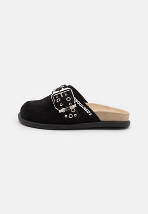 Sandale din suede negru cu talpă plată, având o curea lata cu un cataramă argintie și elemente decorative, completată cu o talpă interioară béj căptușită.