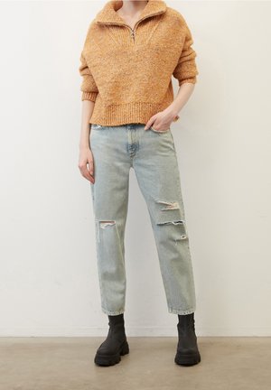 Personne portant un pull zippé orange texturé, un jean bleu clair déchiré et des bottes noires épaisses, debout contre un mur blanc uni.