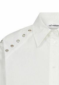 Camicia bianca in cotone con colletto, chiusura a bottoni e dettagli in metallo sulle spalle. Il tessuto è liscio e ha una finitura pulita.