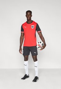 Ερυθρό και μαύρο αθλητικό φανέλα με κοντά μανίκια, σετ με μαύρο σορτς και λευκές κάλτσες. Ο παίκτης κρατάει μια μαύρη και λευκή ποδοσφαιρική μπάλα.