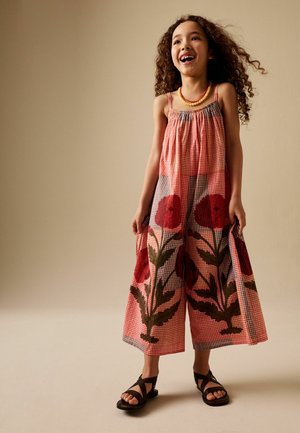 Mädchen mit lockigem Haar, das ein rotes kariertes Kleid mit großen Blumenmustern, schwarze Sandalen und eine gelbe Perlenkette trägt, lächelnd und stehend.