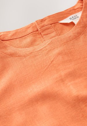 Oranssi pellavapusero, jossa pyöreä pääntie ja näkyvät ompelusaumat. Pehmeä rakenne, luonnollinen ja hieman rypistynyt ulkonäkö.