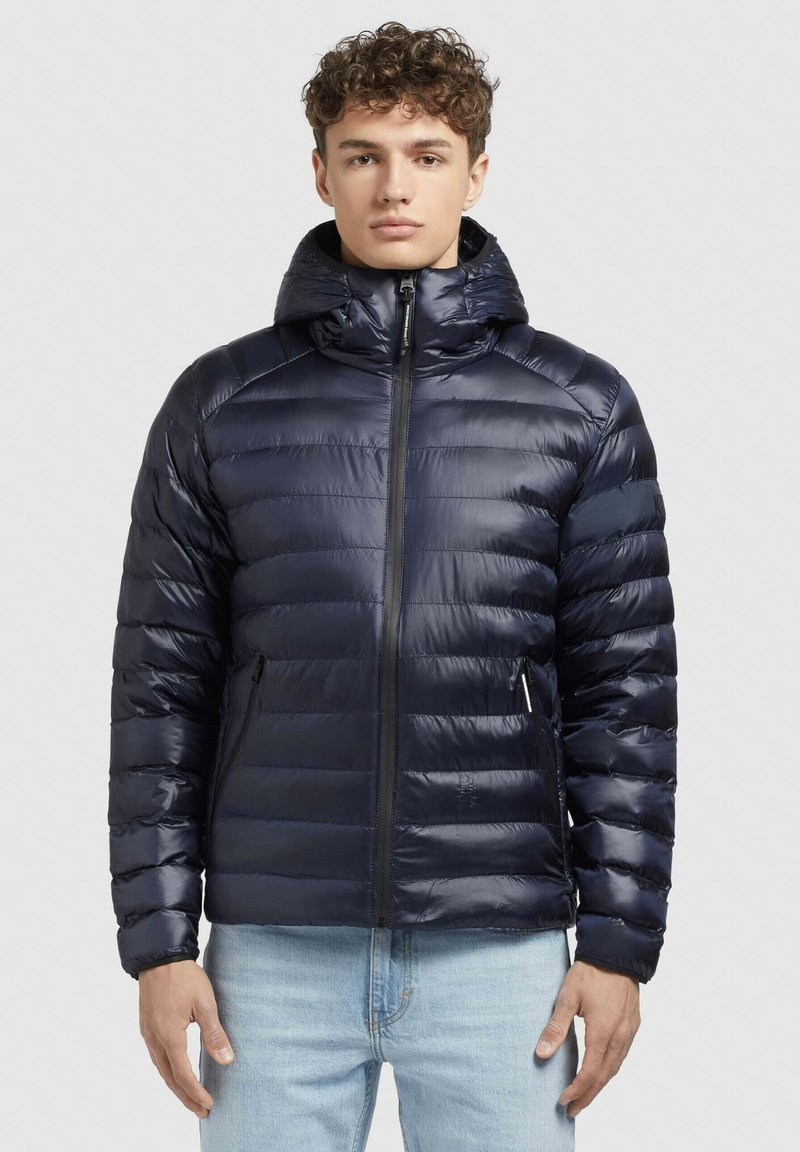 Mornarskoplava pufasta jakna od sjajnog materijala s kapuljačom, s patentnim zatvaračem i dva prednja džepa, dizajnirana s quilted sekcijama.
