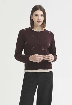 Femme aux cheveux bruns et lisses porte un cardigan bordeaux boutonné avec une broderie florale, un haut beige en dessous et un pantalon sombre à fines rayures.