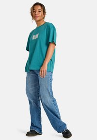 Teal oversized t-shirt with white text, paired with light blue flared jeans and black shoes. Simple design, cotton material, relaxed fit.