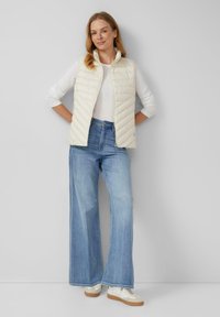 Lichtgewicht, crèmekleurige gewatteerde vest met hoge kraag, gedragen over een wit langemouwen shirt. Gecombineerd met brede blauwe jeans en crèmekleurige sneakers.