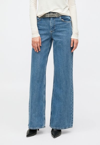 Wijde blauwe spijkerbroek van denim, met een hoge taille, vijf zakken en subtiele vervaging. Gecombineerd met een zwarte riem met zilveren accenten en hakken.