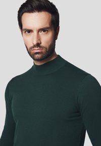 Ciemnozielony sweter z wysokim kołnierzem, prążkowany, o dopasowanym kroju i gładkiej fakturze, z długimi rękawami i przylegającą sylwetką.
