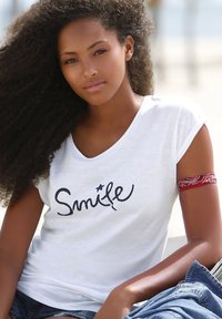 Camiseta blanca de manga corta con la palabra "Sonríe" impresa en cursiva negra, combinada con pantalones cortos de denim azul y una banda roja estampada en el brazo superior.