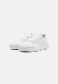 Des baskets en cuir blanc présentent un bout arrondi, des détails perforés, des lacets plats et une semelle plateforme avec un logo discret sur la languette.