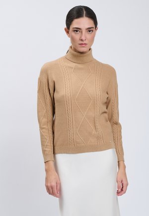 Kvinde med mørkt hår bundet tilbage iført en beige kabelstrikket rullekrave og et hvidt skørt, stående mod en ensfarvet lys baggrund.