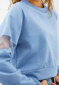 Světle modrý svetr vyrobený z měkké tkaniny s texturovaným povrchem. Obsahuje síťovinové detaily na rukávech a má volný střih.