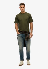 Olivengrøn bomulds T-shirt med korte ærmer, parret med lyseblå jeans og en ternet flannel skjorte bundet om livet. Hvide sneakers.