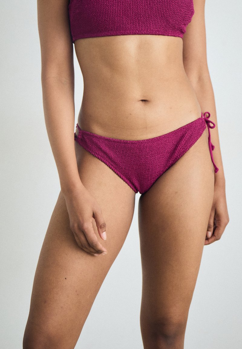 Fuksijos spalvos dviejų dalių bikini, tekstūruotas mezgimo audinys. Viršus yra bandeau stiliaus, o apačioje yra reguliuojami raišteliai šonuose.