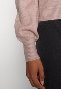 Jasnoróżowy sweter z dzianiny z prążkowanymi mankietami, charakteryzujący się miękką teksturą. Rękaw ma puszysty fason, w zestawieniu z ciemnym dżinsem.