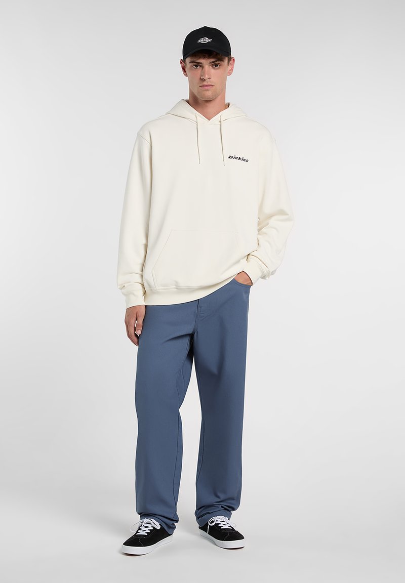 Crèmekleurige hoodie met voorzak en zwart logo; blauwe broek met een relaxte pasvorm; zwarte pet; zwarte schoenen met witte veters.