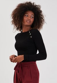 Top noir à manches longues côtelé avec quatre boutons dorés sur l'épaule gauche, associé à un pantalon bordeaux taille haute.