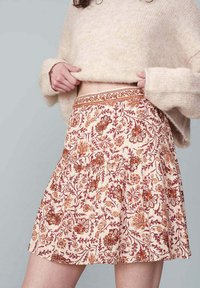 Jupe à motif floral en crème, avec des dessins botaniques rouges et orangés. Un tissu texturé avec une taille froncée et une forme évasée.