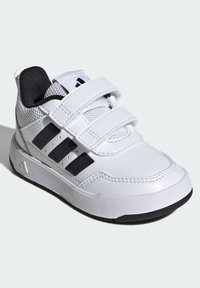 Bijela sportska cipela s dvije crne Velcro trake, teksturiranim gornjim dijelom od sintetičkog materijala i gumenim potplatom. Na boku su tri crne pruge.