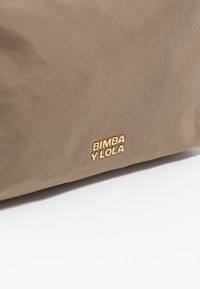 Beżowa torba materiałowa o gładkiej fakturze, z wytłoczonym złotym logo "BIMBA Y LOLA" umieszczonym w prawym dolnym rogu.