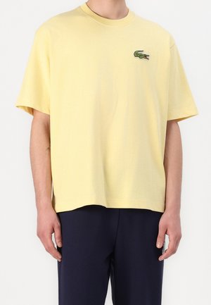 Camiseta de algodón amarilla con cuello redondo y mangas cortas. Presenta un logo de cocodrilo verde bordado en el lado izquierdo del pecho.