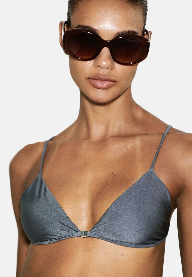 Femme portant des lunettes de soleil oversize marron foncé et un haut de bikini en satin gris à fines bretelles, sur un fond uni.