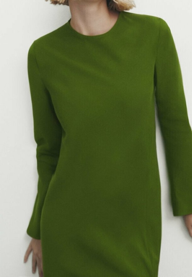 Femme portant une robe longue à manches longues, longueur genou, couleur vert olive, se tenant devant un fond clair uni.